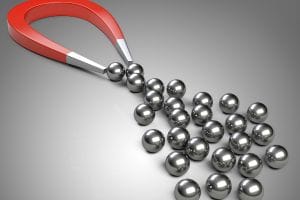 A red horseshoe magnet attracts several shiny metallic spheres, forming a chain, with more spheres scattered on a grey surface—illustrating how you can attract the right staff members to build a strong, connected team to grow your accounting firm.
