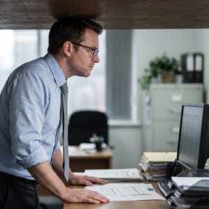 A man in business attire stands at a desk, leaning forward to look at a computer monitor, and his head is touching the seiling. This represents accountancy firm owners hitting a "plateau" or "ceiling".